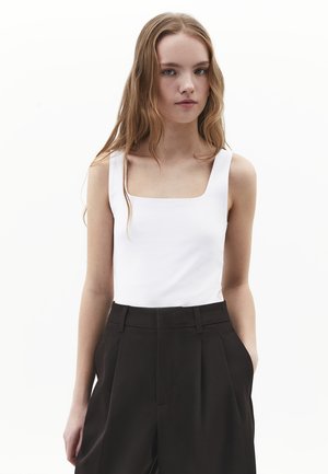 Giovane donna con lunghi capelli castano chiaro che indossa una canottiera bianca senza maniche con scollo quadrato e pantaloni neri plissettati a vita alta.