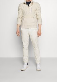 Pullover in pile crema con mezza zip, dotato di tasca sul petto e polsini elasticizzati, abbinato a pantaloni tapered crema e sneakers bianche.