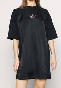 Žena v černých oversized šatech s krátkým rukávem, s lesklým panelem na přední straně a malou vícebarevnou květinovou emblémem na hrudi.