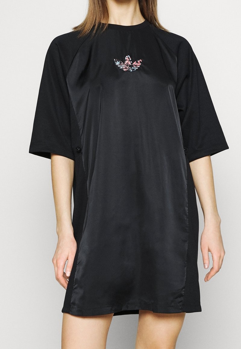 Žena v černých oversized šatech s krátkým rukávem, s lesklým panelem na přední straně a malou vícebarevnou květinovou emblémem na hrudi.