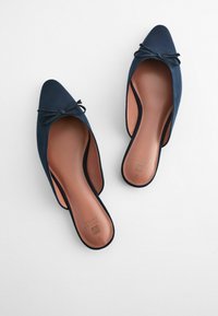Mules en bleu marine à bout pointu avec une texture lisse, présentant un détail en nœud sur le devant et des semelles beiges avec un logo.