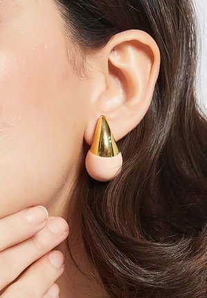 Guld- og blushfarvede øreringe med en konusform på toppen af en rund base. Glat metal og matte overflader er synlige.