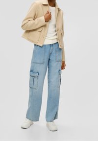 Giacca cropped beige con tasche, indossata sopra un maglione bianco in maglia e jeans cargo a gamba larga blu chiaro con tasche laterali. Sneakers bianche.