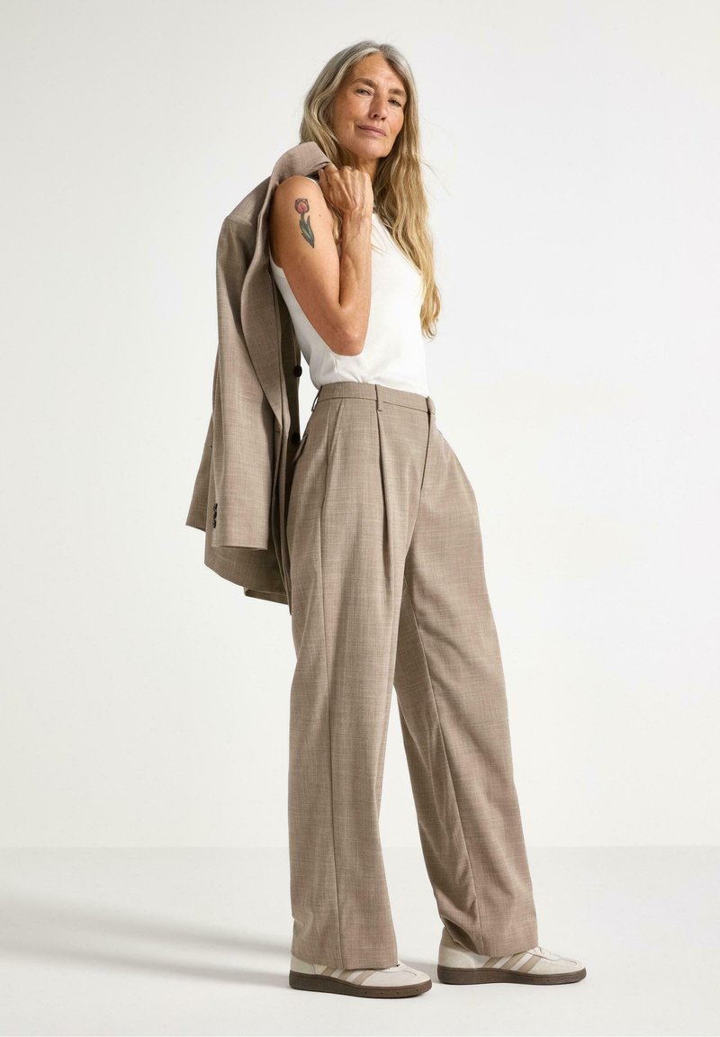 Lys tan, teksturerede bukser med folder; parret med en hvid tanktop. En grå blazer hviler på skulderen. Accentueret med cremefarvede sko.