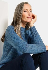 Maglione blu lavorato a maglia caratterizzato da una consistenza spessa e polsini a costine, abbinato a pantaloni scuri. L'illuminazione mette in risalto i dettagli del tessuto.