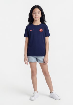 Jente med langt mørkt hår iført en mørkeblå Nike T-skjorte, lyse denimshorts og hvite sneakers, stående foran en hvit bakgrunn.