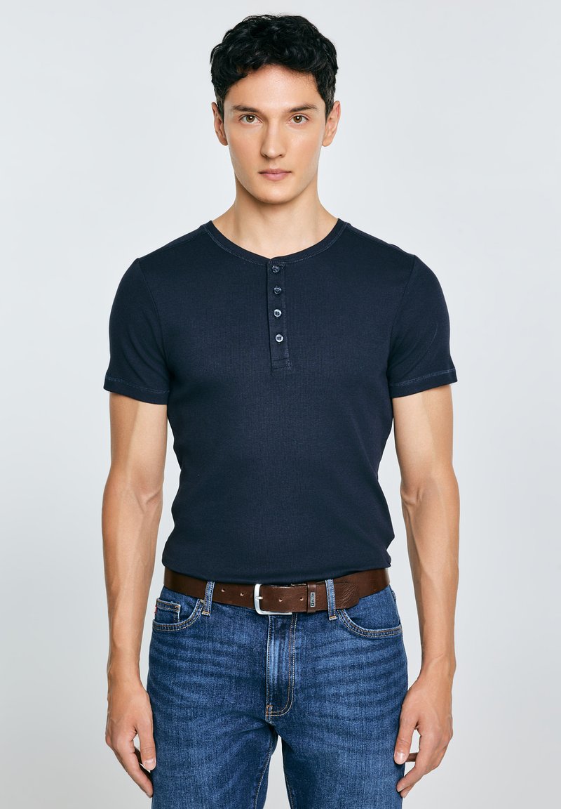 Mand iført sort henley-shirt med korte ærmer, stoppet ned i blå jeans med brunt bælte, stående mod en ensartet grå baggrund og kigger fremad.