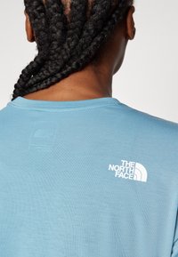 Heled sinine T-särk, mis on valmistatud pehmest kangast, featuring valge logo "The North Face" ja ruudukujuline silt seljal kaela lähedal.