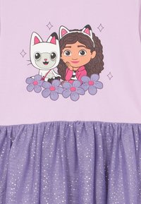 Deklica z glavnikom v obliki mačjih ušes sedi ob belem mačku s podobnimi uši, obdana s vijoličnimi cvetovi v roza in vijoličasti iskrivi obleki.