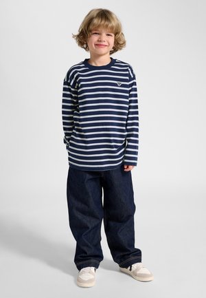 Młody chłopiec stoi, uśmiecha się, ubrany w granatowo-biały pasiasty longsleeve, ciemne szerokie dżinsy oraz białe trampki na jednolitym tle.