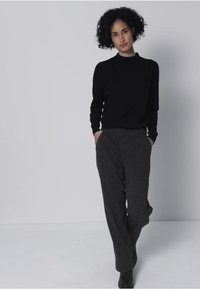 Pull noir ajusté à manches longues, associé à un pantalon large gris foncé. Le modèle est debout, les mains dans les poches, devant un fond uni.