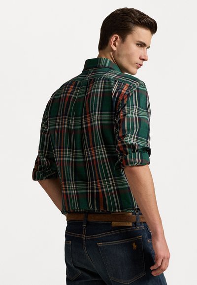 Camisa a cuadros verde y azul marino con mangas arremangadas, que presenta franjas rojas y naranjas. Combinada con jeans azul oscuro y un cinturón marrón.