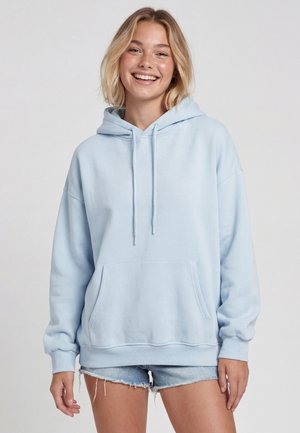 Młoda kobieta uśmiecha się, ubrana w jasnoniebieską oversize bluzę z kapturem i postrzępione szorty jeansowe, stoi na tle jednolitego jasnego tła.