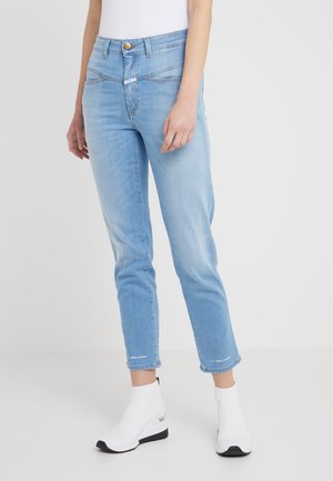 Jasnoniebieskie jeansy do kostek z unikalnymi ukośnymi przednimi kieszeniami, noszone z białymi botkami przypominającymi skarpetki na jednolitym tle.