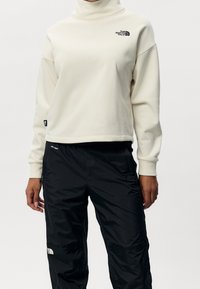 Kermanvärinen poolokauluksinen collegepaita, jossa musta logo, yhdistetty mustiin, kevyisiin, vedenpitäviin housuihin, joissa on vetoketjut ja taskut.