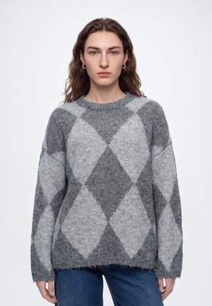 Personne aux longs cheveux bruns portant des boucles d'oreilles créoles dorées et un pull gris à motif losanges, debout devant un fond blanc uni.
