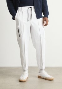 Λευκά cargo παντελόνια σε κοφτή γραμμή με ελαστική μέση και κορδόνι, διαθέτοντας πλαϊνές τσέπες με φερμουάρ. Συνδυασμένα με λευκά αθλητικά παπούτσια.