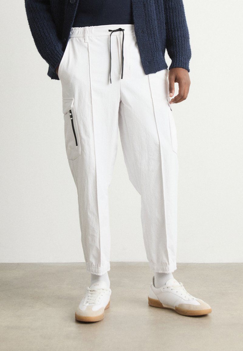 Λευκά cargo παντελόνια σε κοφτή γραμμή με ελαστική μέση και κορδόνι, διαθέτοντας πλαϊνές τσέπες με φερμουά ρ. Συνδυασμένα με λευκά αθλητικά παπούτσια.