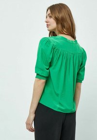 Blusa verde a maniche corte con tessuto strutturato, schiena arricciata e polsini delle maniche arricciati, abbinata a pantaloni neri.