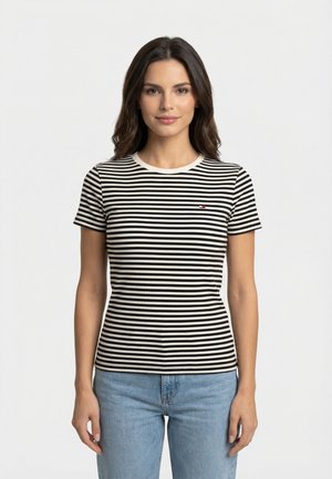 Donna con maglietta a righe orizzontali bianche e nere e jeans azzurri, in piedi davanti a uno sfondo bianco semplice.