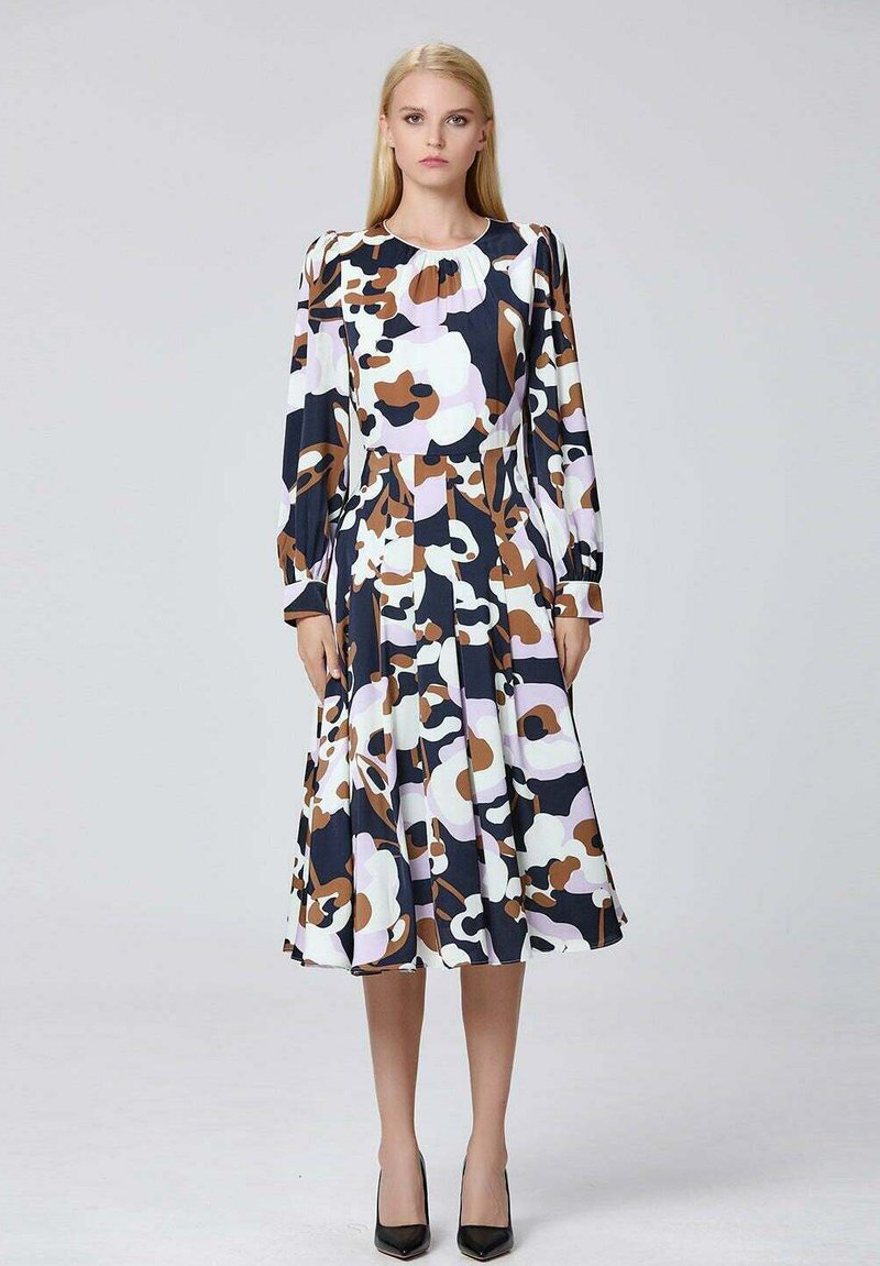 Femme se tenant debout dans une robe à manches longues, longueur genou, avec des motifs floraux abstraits noirs, bruns, blancs et lavande, portant des talons noirs.