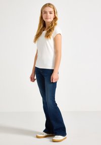 Hvit t-skjorte med korte ermer, mørkeblå flared jeans og hvite sneakers med brune såler. Modellen står mot en nøytral bakgrunn.