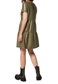 Robe en simili cuir vert olive, à manches courtes, avec un design à volants et un dos en forme de trou de serrure. Associée à des bottines noires à semelles épaisses.