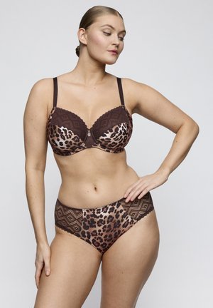 Frau posiert in brauner und leopardenmusternder Lingerie mit Spitzen-Details, stehend vor einem schlichten hellgrauen Hintergrund.