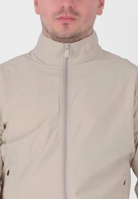 Giacca leggera beige con colletto alto e zip a tutta lunghezza. Presenta una texture liscia e opaca e dettagli di cucitura minimi.