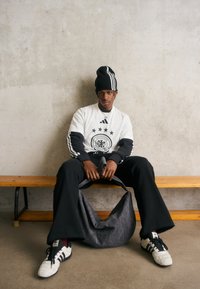 Svart lue med hvite striper, hvit langermet skjorte med svarte detaljer og logo, svarte bukser, og en grå veske på en benk.