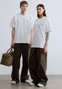 Λευκά βαμβακερά μπλουζάκια με ένα μικρό πράσινο λογότυπο, συνδυασμένα με καφέ φαρδιά παντελόνια. Μια μπεζ τσάντα κρατιέται σε ένα χέρι, με καφέ διακοσμητικά στοιχεία.