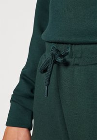 Dark green sweatshirt fabric with a textured finish. Features an adjustable waistband with a braided drawstring and metal aglets.