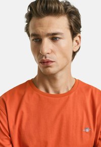 Orange bomulds t-shirt med rund hals og korte ærmer, med et lille broderet logo på venstre bryst. Glat tekstur.
