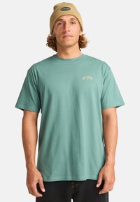 Camiseta de algodón verde menta con mangas cortas y escote redondo. Presenta un pequeño logo beige en el pecho. Se lleva con pantalones negros.