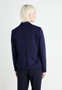Blazer aderente blu navy con spalle strutturate, tessuto liscio, cucitura posteriore unica e design minimalista. Abbinato a pantaloni scuri.