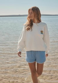 Sweatshirt blanc surdimensionné avec un design brodé. Associé à un short à rayures bleues et blanches avec un ourlet festonné, se tenant dans l'eau peu profonde.