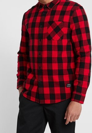 Camisa de franela a cuadros rojos y negros con botones en la parte delantera, mangas largas y un bolsillo en el pecho. La tela parece suave y texturizada. Etiqueta negra en el dobladillo.