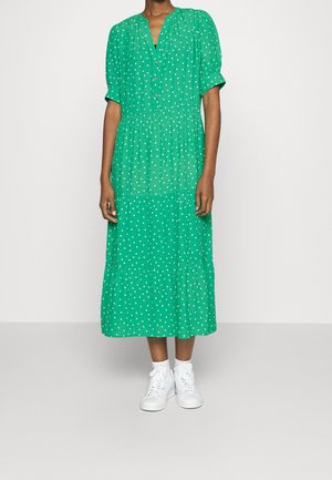 Zielona sukienka midi w białe kropki z krótkimi rękawami i zapinanym na guziki dekoltem. Noszona z białymi trampkami dla kontrastu. Lekka tkanina, warstwowy fason.