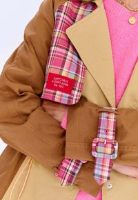 Béžový trenčkot s ružovým károvaným podšívkou, vybavený výraznou prackou a farebným šitím. Červený štítok hovorí: "ŠŤASTIE TI DOBRE SEDÍ."