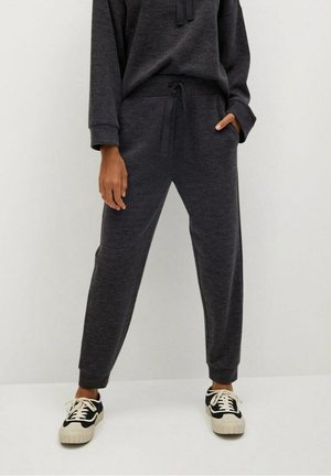 Personne portant un pantalon de survêtement gris foncé avec cordon de serrage et un sweat assorti, debout avec une main dans la poche et des baskets noires et blanches.