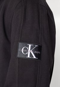 Sudadera de algodón negra con textura acanalada, cuello redondo y una etiqueta negra prominente con texto blanco que dice "Calvin Klein Jeans" y "CK."