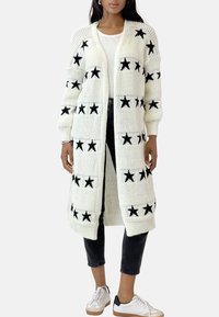 Vit stickad cardigan med svarta stjärnmönster, öppen fram, drop shoulders och ribbade ärmslut. Matchad med mörka jeans och vita sneakers.
