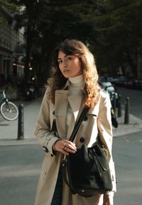 Beiger Trenchcoat mit schwarzen Knöpfen, getragen über einem weißen Rollkragenpullover. Hält eine schwarze Wildleder-Bucket-Bag mit Zugbandverschluss und goldenen Akzenten.