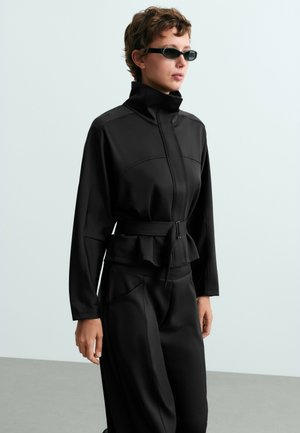 Personne portant une veste noire à col montant avec ceinture et un pantalon noir, portant également des lunettes de soleil rectangulaires noires, debout devant un fond uni.