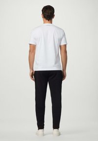Hombre de pie de espaldas a la cámara, vistiendo una camiseta blanca, pantalones de chándal negros y zapatillas blancas, frente a un fondo simple.