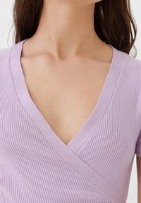 Kvinna i en ribbad, lavendel V-ringad omslagstopp som visar halslinjen och övre bröstet, med långt brunt hår.