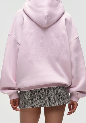 Osoba w jasnoróżowej, oversize'owej bluzie z haftowanym napisem na plecach i czarno-białej, wzorzystej mini spódnicy.
