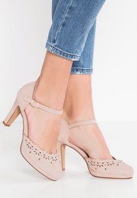 Stopy w jasnobeżowych pantoflach na obcasie typu Mary Jane z kwiatowymi wycięciami, zestawione z przyciętymi niebieskimi dżinsami.