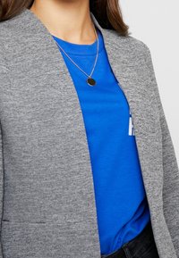 Blazer gris texturé porté sur un T-shirt bleu avec des lettres blanches, collier en argent avec un pendentif circulaire, sur fond neutre.