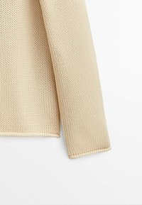 Manche de pull en tricot beige avec un motif de points texturé et des bords soigneusement finis, sur fond blanc.